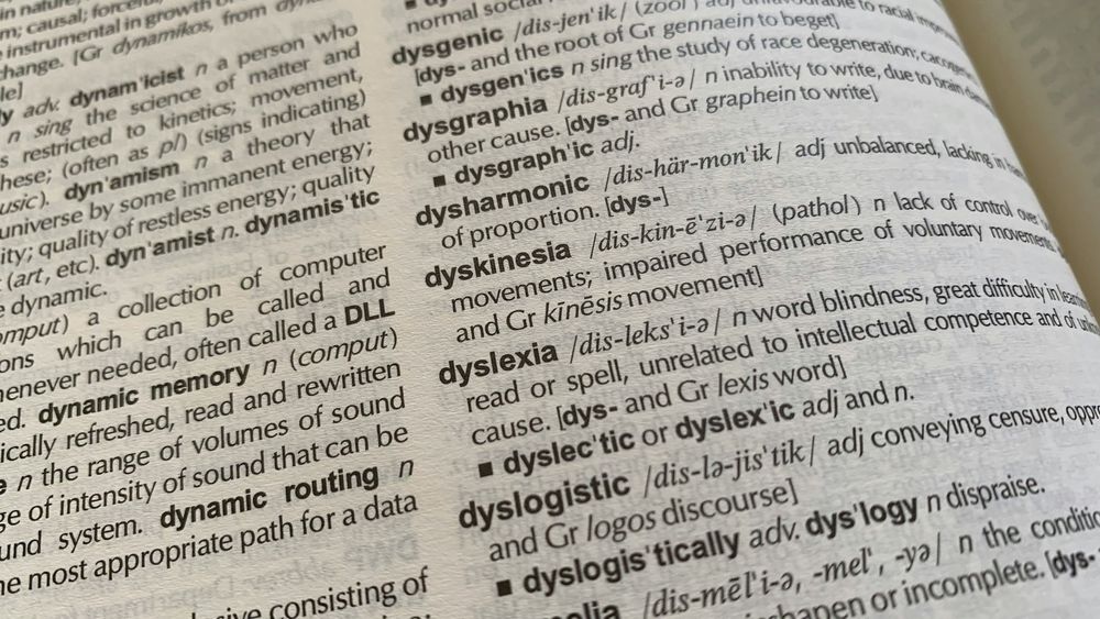 Un gros plan sur la page d'un dictionnaire ouvert. L'entrée principale visible et mise en évidence est "dyslexia" (dyslexie), avec sa définition : "a word blindness, great difficulty in learning to read or spell, unrelated to intellectual competence and of unknown cause."