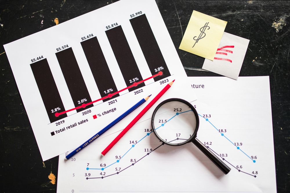 Deux graphiques d'analyse de données sur un bureau noir, accompagnés de crayons rouge et bleu, d'une loupe et d'une note autocollante avec un signe dollar, illustrant l'audit et l'analyse des coûts et revenus liés à l'accessibilité d'un site web.